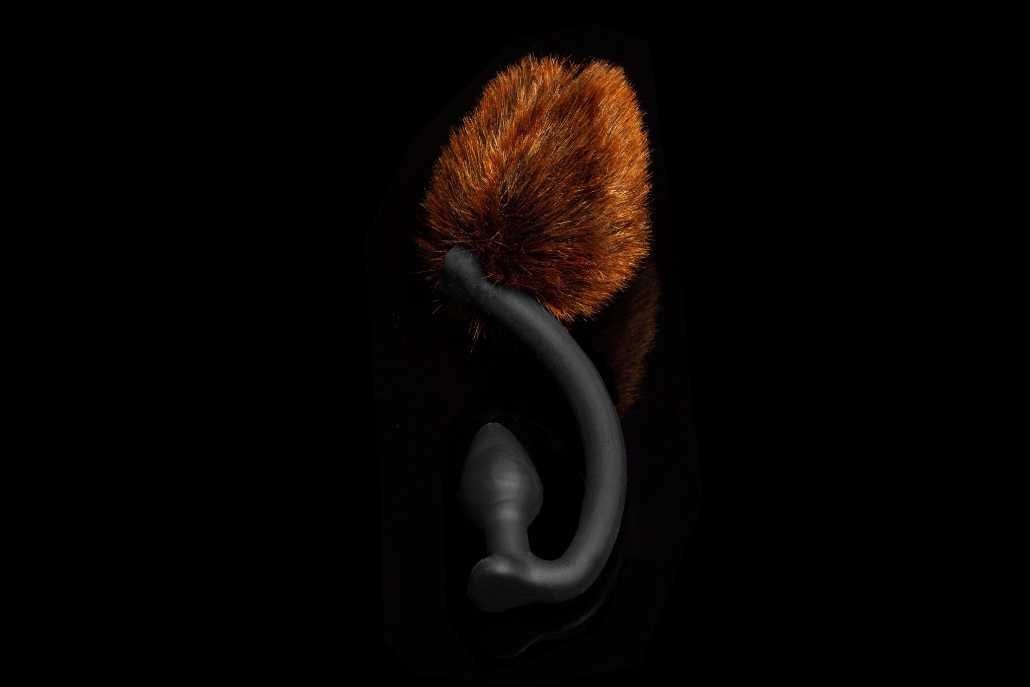 Brown Bear tail butt plug made of silicone and faux fur, but the plug is hooked so when worn, the tail sits at the bottom of the lumbar area. It’s laying flat on a black background.