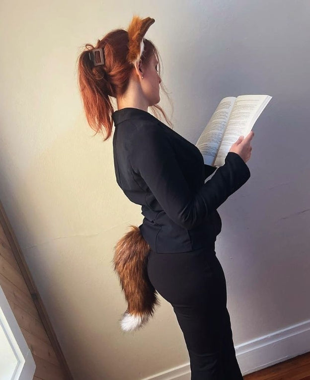 Person wearing a black outfit with a fox tail and ears, reading a book against a plain wall.