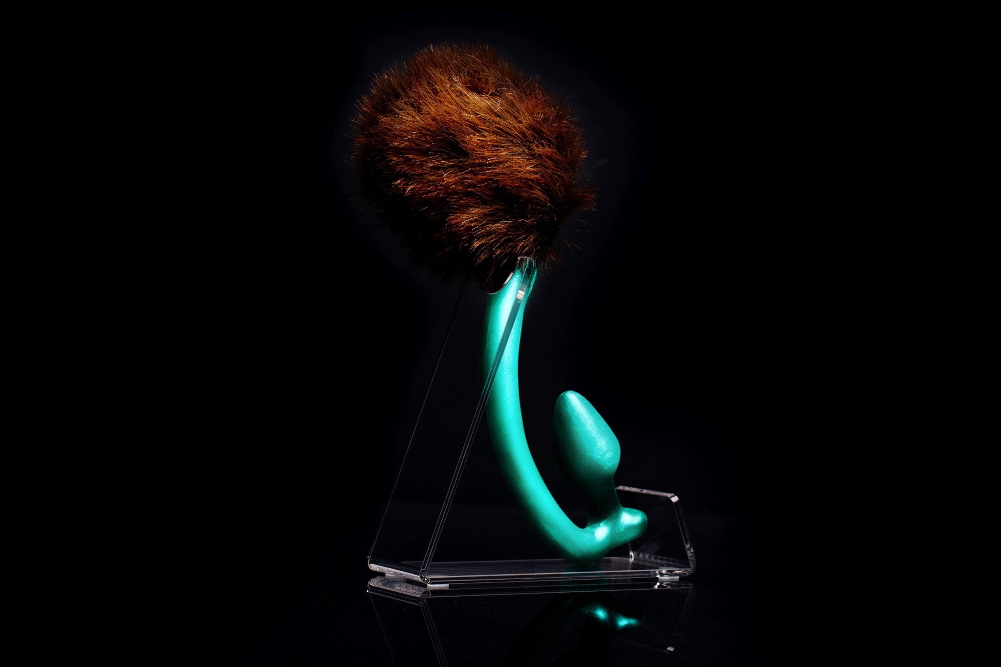 Brown Bear tail butt plug made of silicone and faux fur, but the plug is hooked so when worn, the tail sits at the bottom of the lumbar area. It’s standing up on a transparent stand against an all black background.