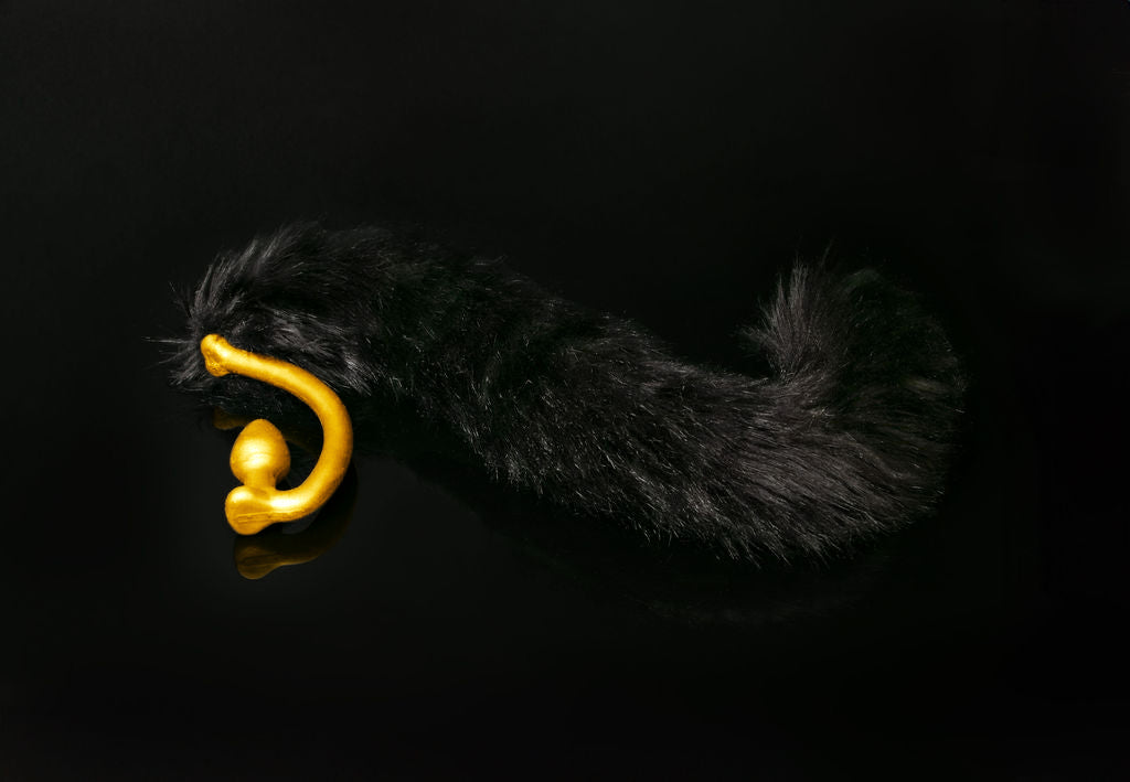Long hair, black cat tail butt plug made of silicone and faux fur, but the plug is hooked so when worn, the tail sits at the bottom of the lumbar area. It’s laying flat on a black background.::Gold
