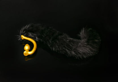 Long hair, black cat tail butt plug made of silicone and faux fur, but the plug is hooked so when worn, the tail sits at the bottom of the lumbar area. It’s laying flat on a black background.::Gold