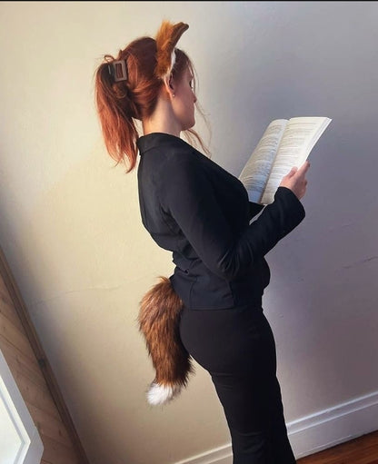 Person wearing a black outfit with a fox tail and ears, reading a book against a plain wall.