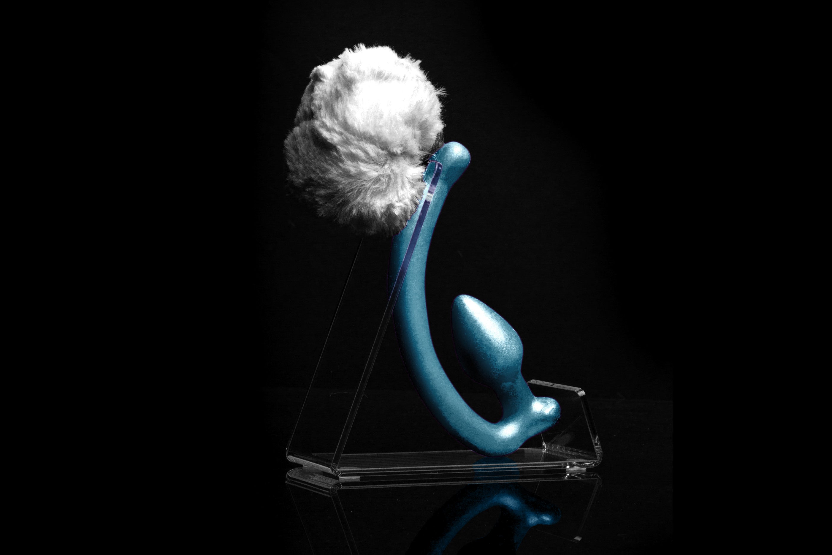 White Rabbit tail butt plug made of silicone and white faux fur, but the plug is hooked so when worn, the tail sits at the bottom of the lumbar area. It’s laying flat on a black background.::Blue