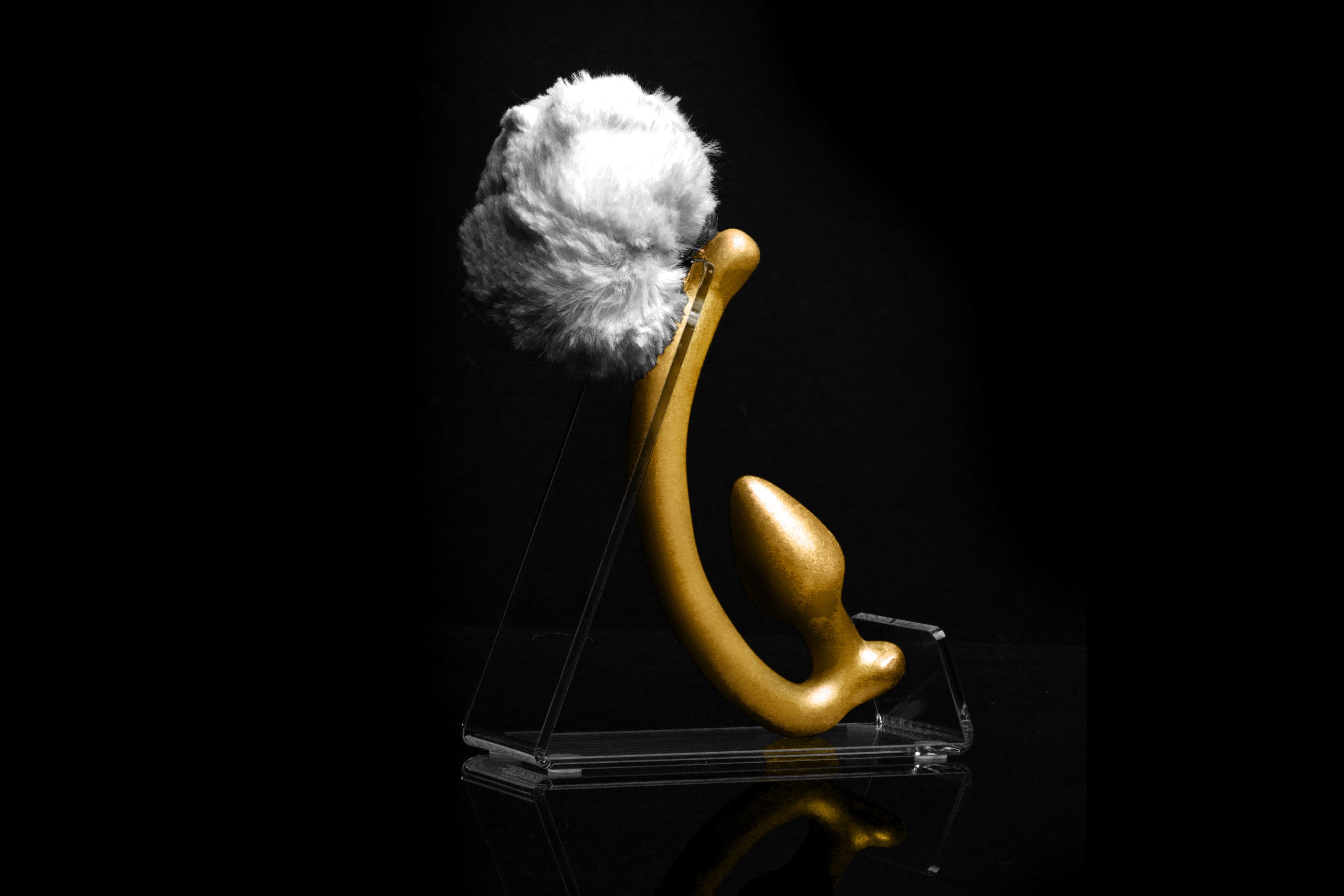 White Rabbit tail butt plug made of silicone and white faux fur, but the plug is hooked so when worn, the tail sits at the bottom of the lumbar area. It’s laying flat on a black background.::Gold