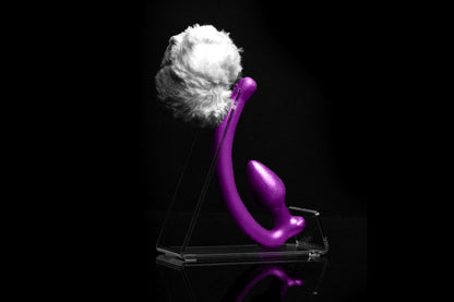 White Rabbit tail butt plug made of silicone and white faux fur, but the plug is hooked so when worn, the tail sits at the bottom of the lumbar area. It’s laying flat on a black background.::Purple