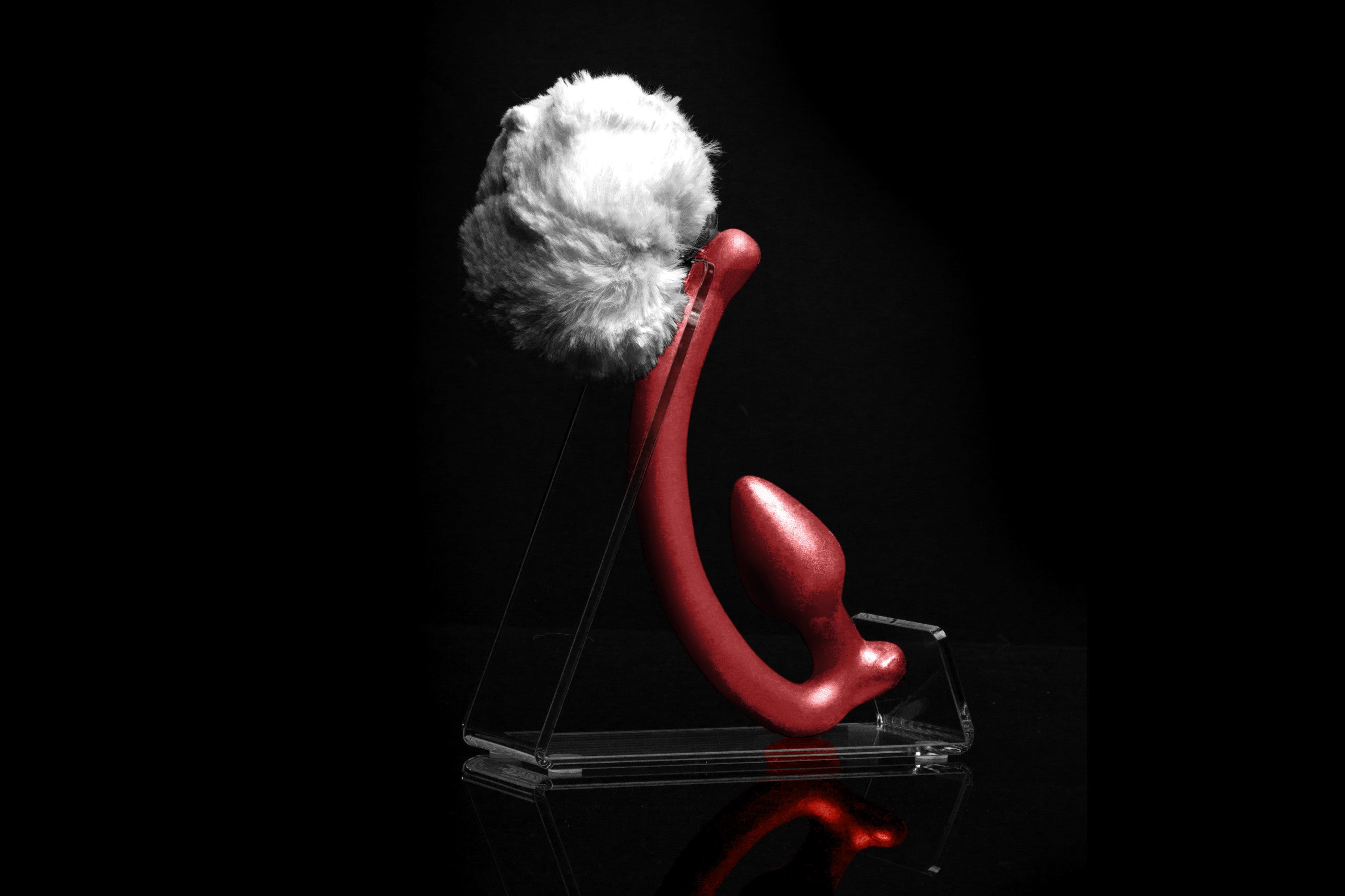 White Rabbit tail butt plug made of silicone and white faux fur, but the plug is hooked so when worn, the tail sits at the bottom of the lumbar area. It’s laying flat on a black background.::Red