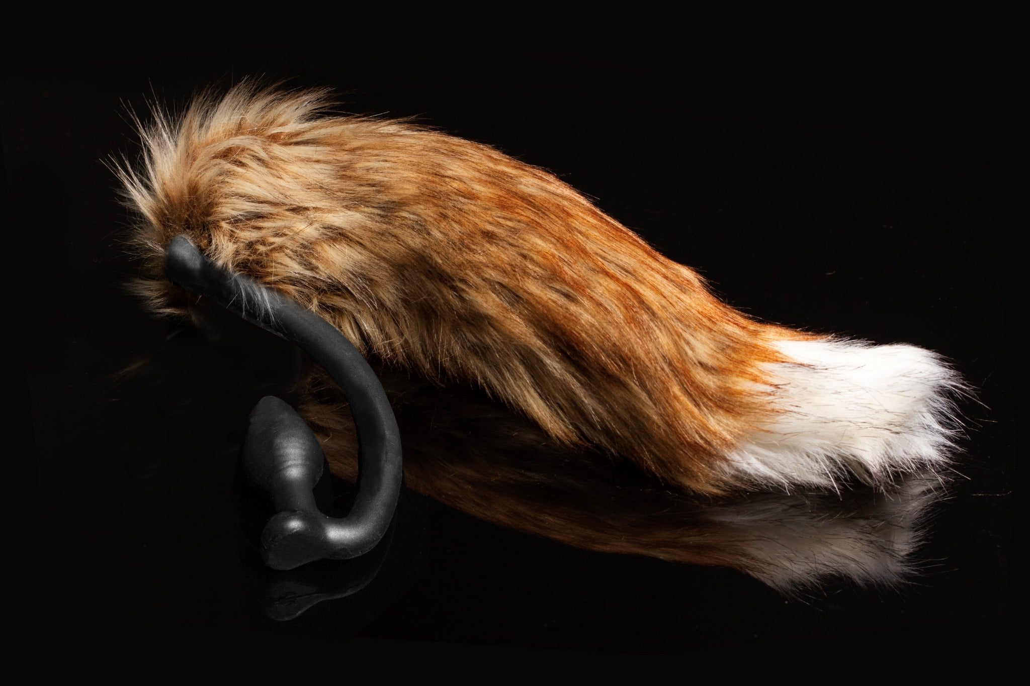 Red Fox tail butt plug made of silicone and faux fur, but the plug is hooked so when worn, the tail sits at the bottom of the lumbar area. It’s laying flat on a black background.