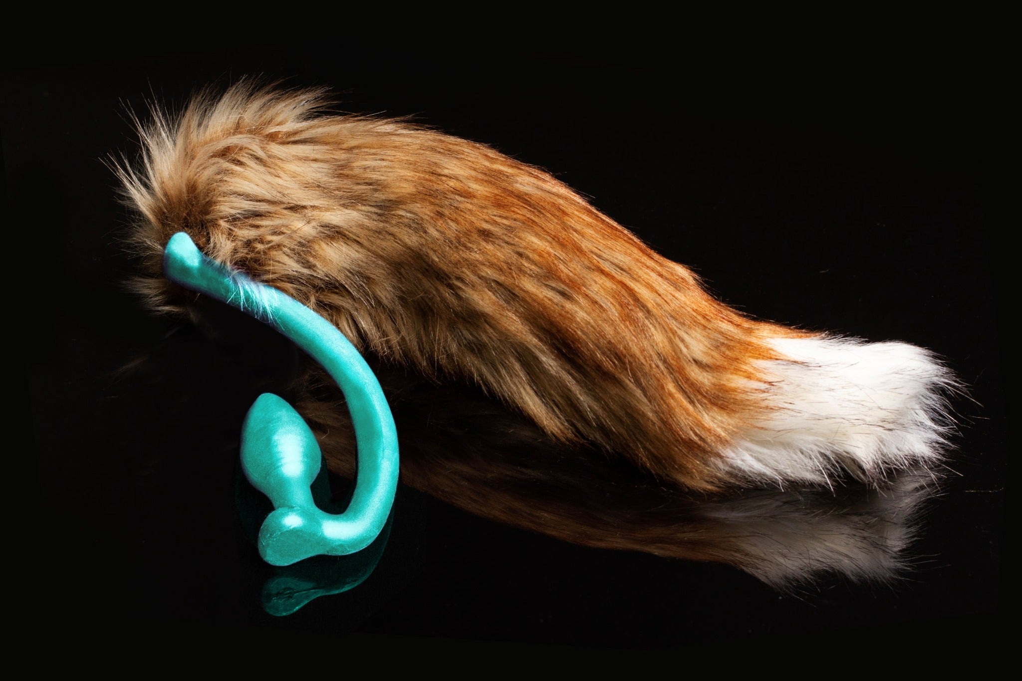 Red Fox tail butt plug made of silicone and faux fur, but the plug is hooked so when worn, the tail sits at the bottom of the lumbar area. It’s laying flat on a black background.