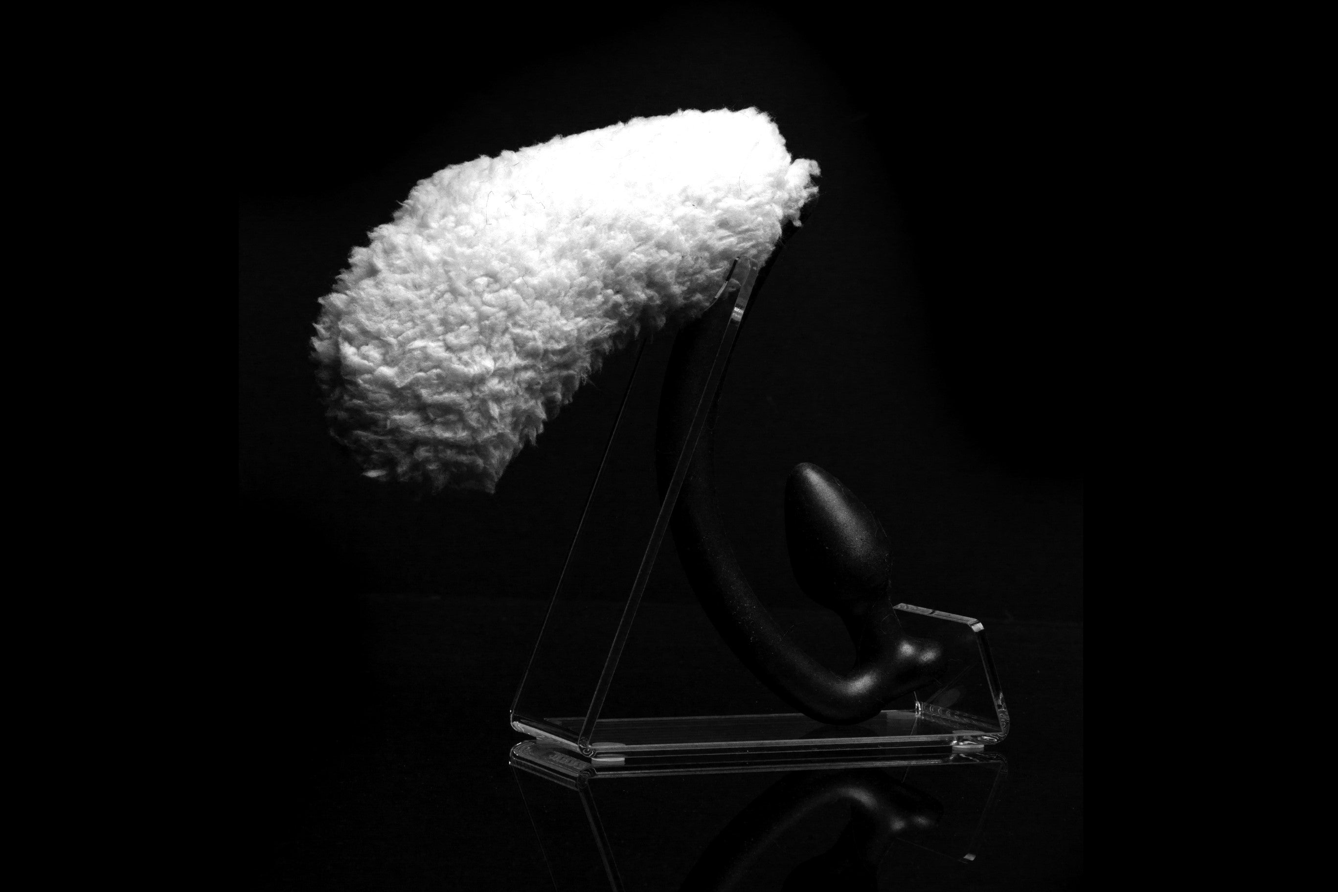 Sheep tail butt plug made of silicone and faux fur, but the plug is hooked so when worn, the tail sits at the bottom of the lumbar area. It’s laying flat on a black background.::Black