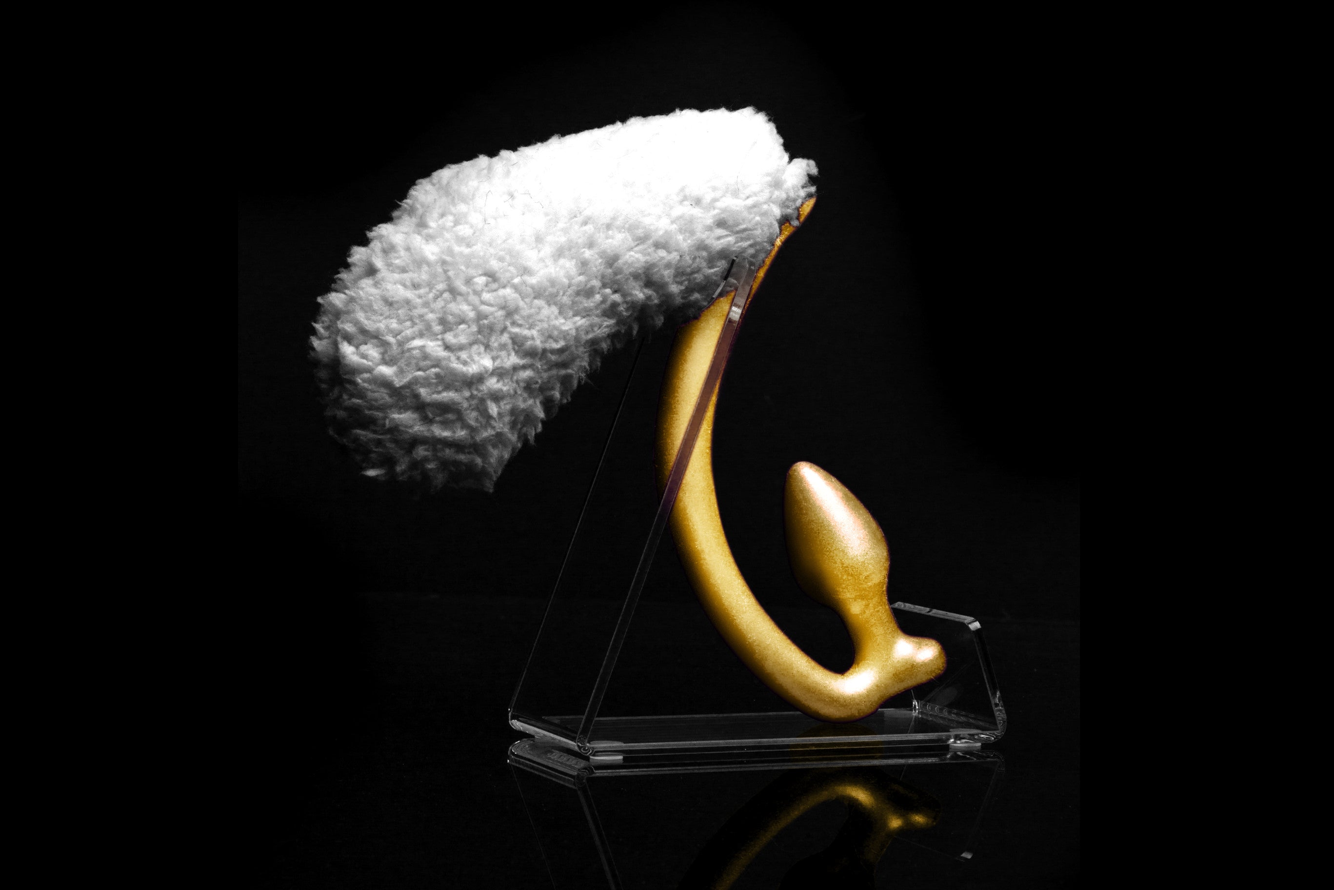 Sheep tail butt plug made of silicone and faux fur, but the plug is hooked so when worn, the tail sits at the bottom of the lumbar area. It’s laying flat on a black background.::Gold