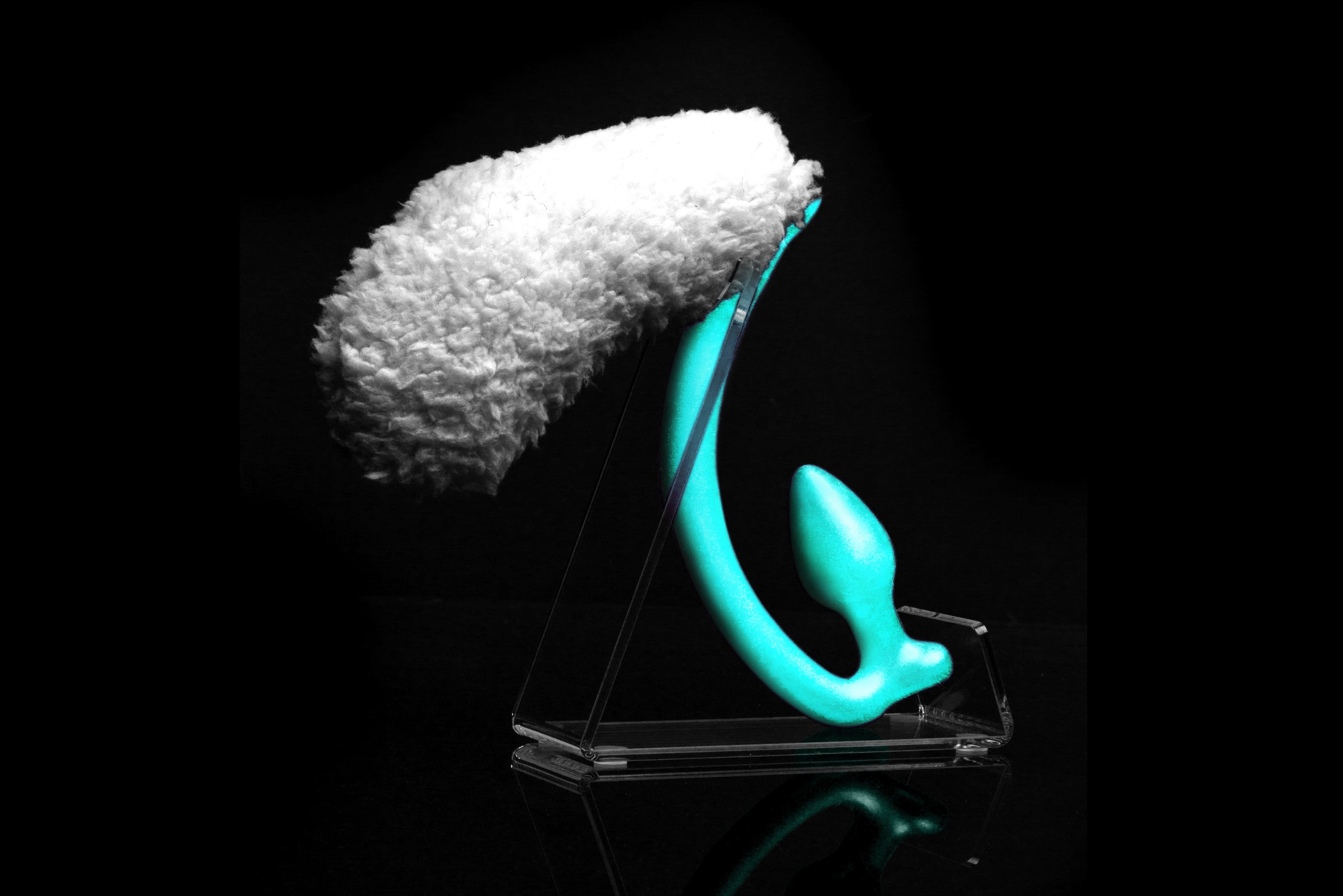 Sheep tail butt plug made of silicone and faux fur, but the plug is hooked so when worn, the tail sits at the bottom of the lumbar area. It’s laying flat on a black background.::Mint