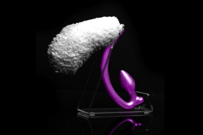 Sheep tail butt plug made of silicone and faux fur, but the plug is hooked so when worn, the tail sits at the bottom of the lumbar area. It’s laying flat on a black background.::Purple