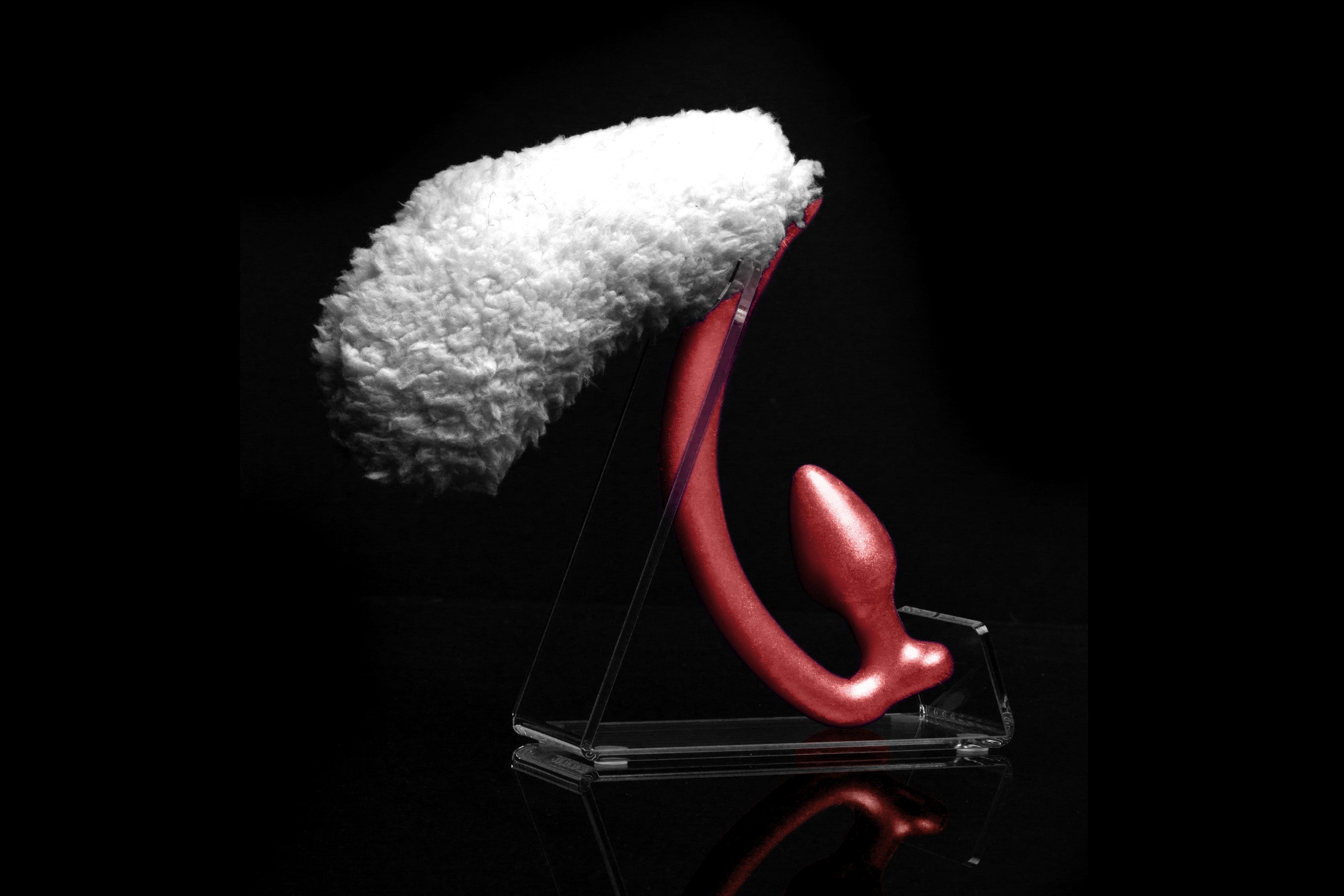Sheep tail butt plug made of silicone and faux fur, but the plug is hooked so when worn, the tail sits at the bottom of the lumbar area. It’s laying flat on a black background.::Red